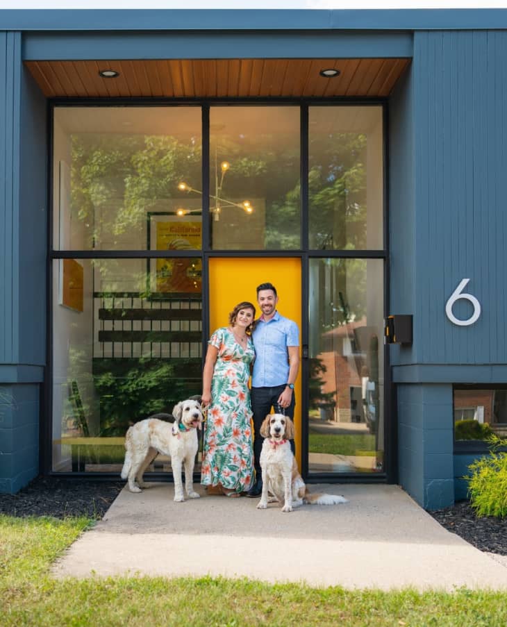 A man and woman standing outside their home with glass floor to ceiling windows with their two dogs