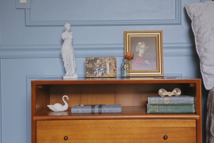 A wooden bedside table with a small statue of a woman and another of a swam with old books and paintings