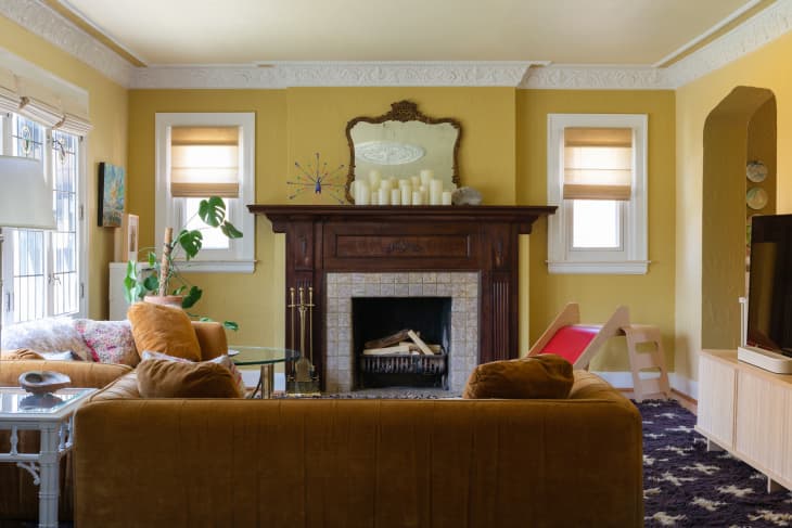 Yellow living room in historic home with vintage mirror above wooden fireplace mantel and intricate crown moulding throughout room.