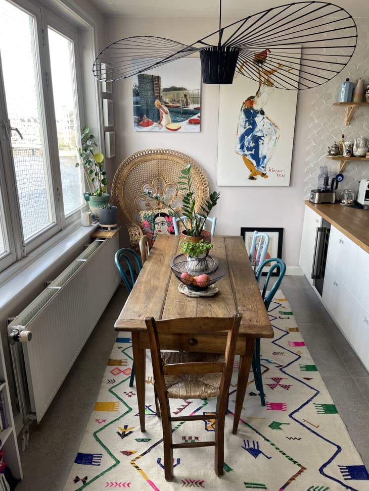Small dining space in apartment with colorful rug on floor and wooden rectangular table surrounded by  mix-matched dining chairs. Large fan pendant light above.