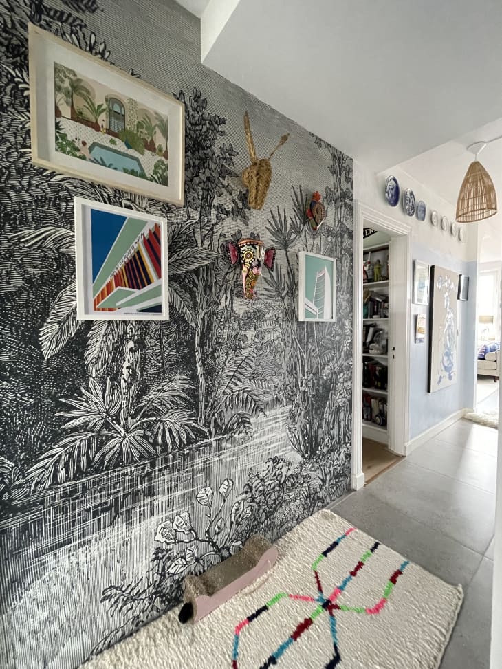 Black, white and grey landscape wallpaper on hallway wall with colorful art prints and a few animal print masks hung on the wall. White rug with colorful accents line the floor.