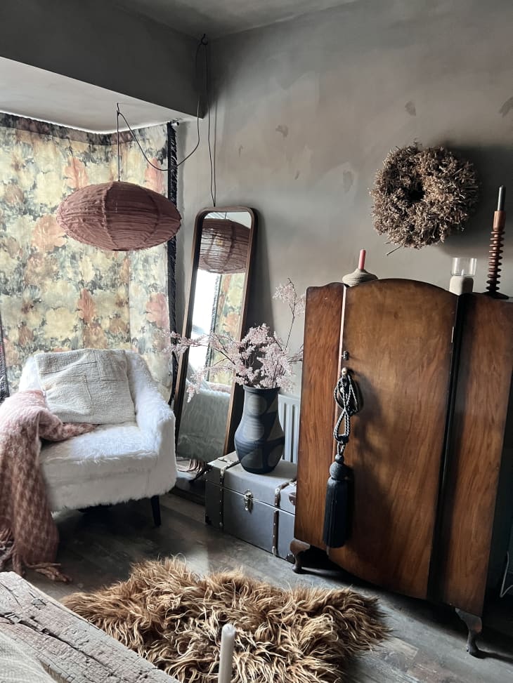 detail of bedroom corner with vintage wood wardrobe, white cushioned accent chair, faux fur shag area rug, full length mirror, rose-colored paper lantern, gray walls