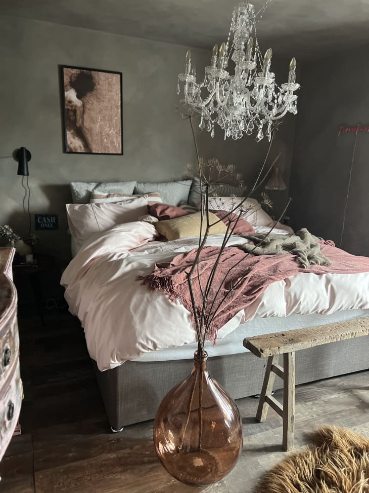 bedroom with gray walls, bed with white and rose colored linens, crystal chandelier, vase with dried branches, rustic wood bench at foot of bed