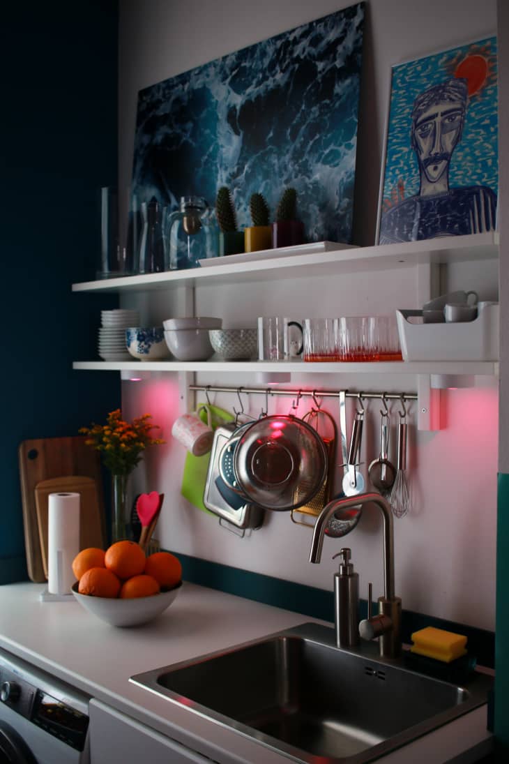 Colorful kitchen with art filled shelving and pink lights under shelves. A bowl of oranges on the countertops.