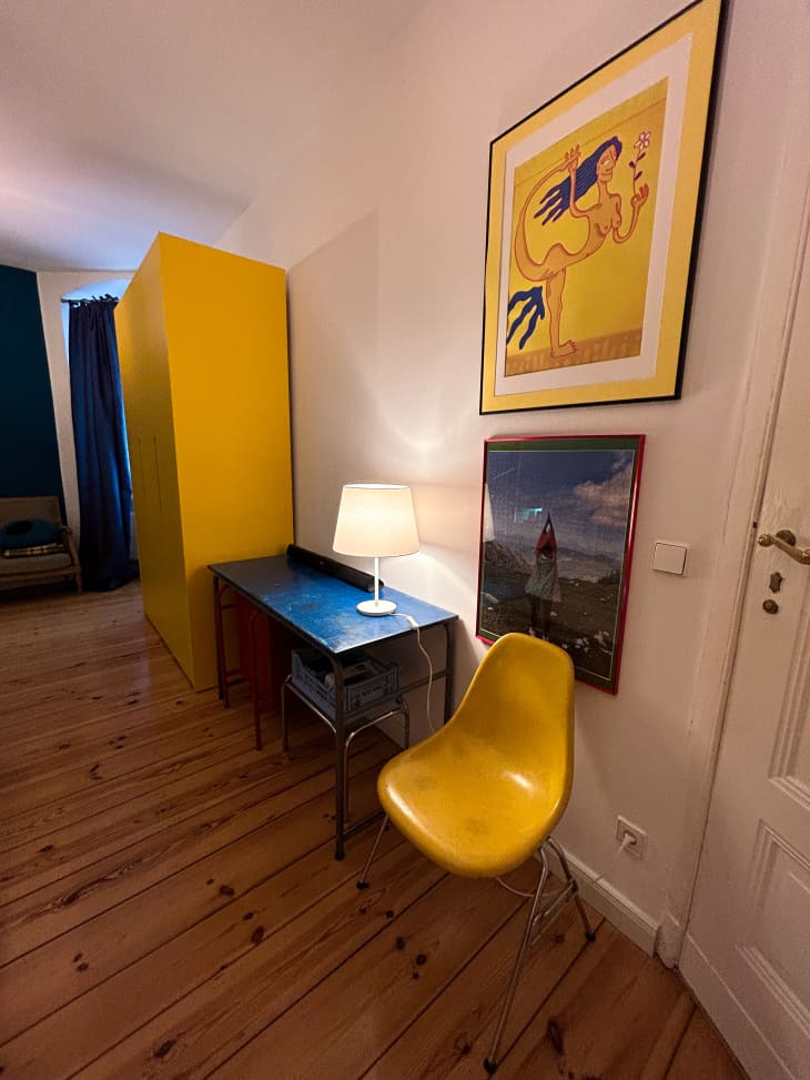 Yellow chair, artwork and storage unit in bedroom with a blue desk with a white lamp on top.
