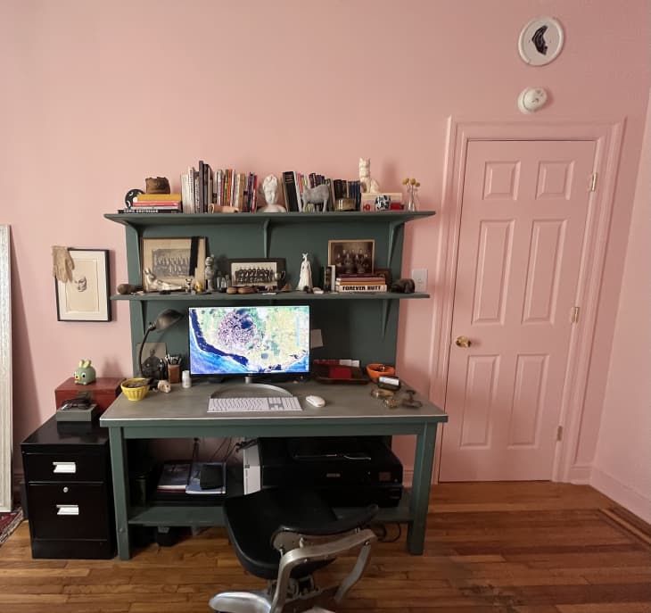 Green desk in home office/workspace. Pink walls, lots of books and art objects on desk shelves