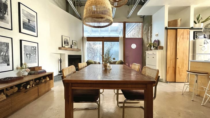 Large dining room with rectangular wood table, rattan-backed dining chairs, natural fiber pendant lamp. Large windows in back of room. the kitchen bar area is visible to the right