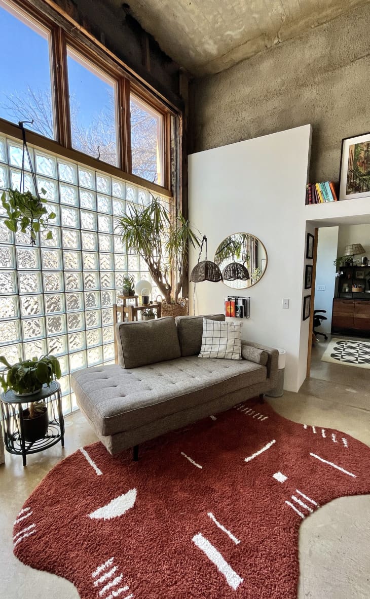 Living room or seating area with gray lounger/chaise, large glass black window with paned windows above, stone finish walls and ceiling, wavy brick red rug with white pattern, lots of plants