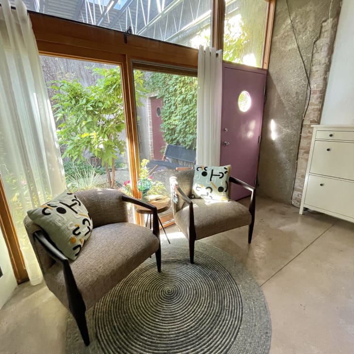 Sitting area with 2 cushioned chairs with patterned throw pillows, large wall of windows behind, white dresser to the right, natural fiber round rug