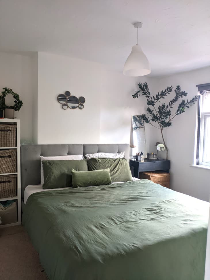 Light filled bedroom with grey upholstered bed frame and bed made with light green linen.  White pendant light hung above bed. Tree branch arrangement in corner of night stand.