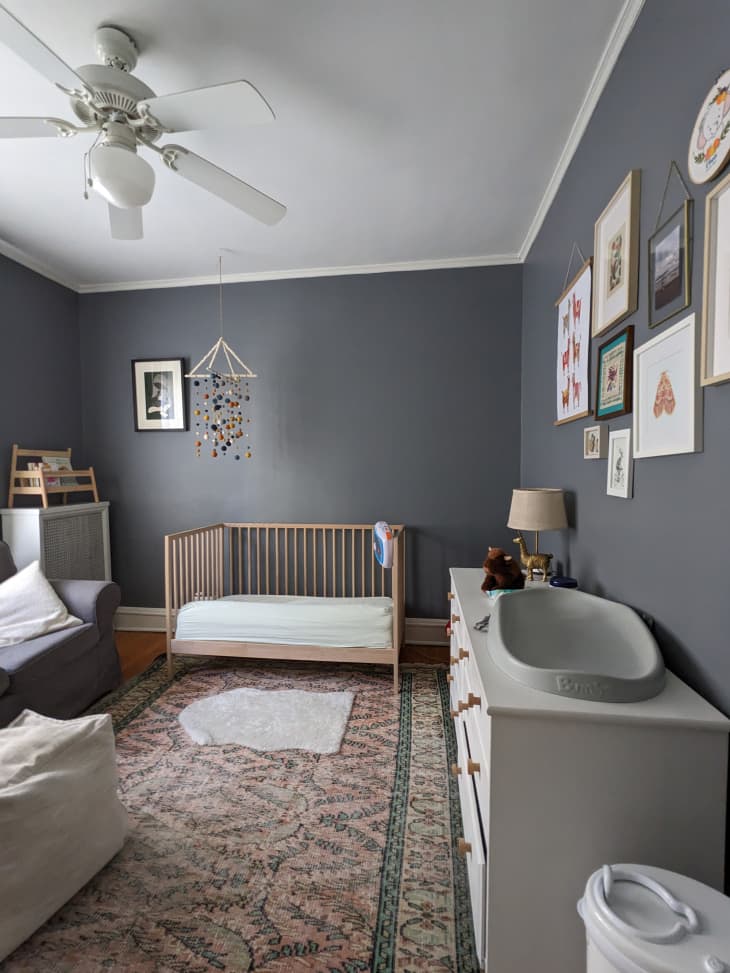nursery with dark gray walls and white ceiling, wood crib, long white dresser, gray armchair, patterned rug
