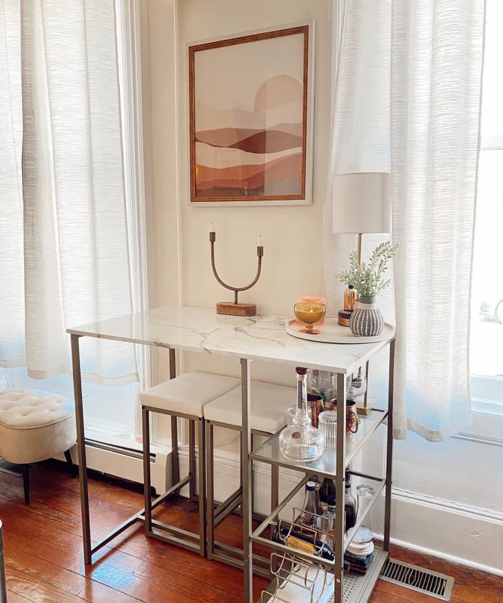 Marble and chrome bar cart with multiple shelves. Candles, lamp on top of cart with art print mounted on wall above.