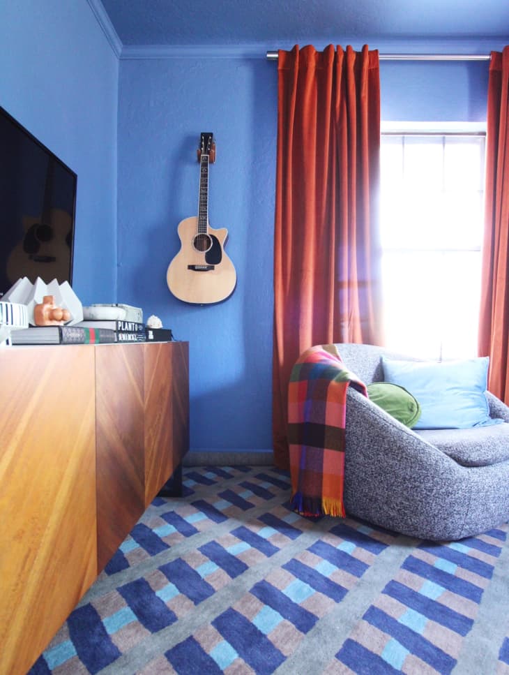 Room with blue walls, guitar hanging on the wall, large wood credenza with TV, cozy blue accent chair, blue and gray geometric rug, rust-colored curtains
