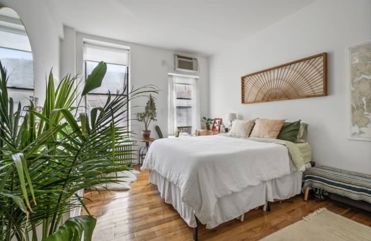 Bedroom with white walls, lots of plants, bed with white linens with natural and green accents, wood floor
