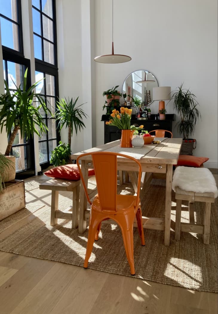 Wooden dining table in sunny room with benches on either side of the table and a orange aluminum dining chair at the head of the table.