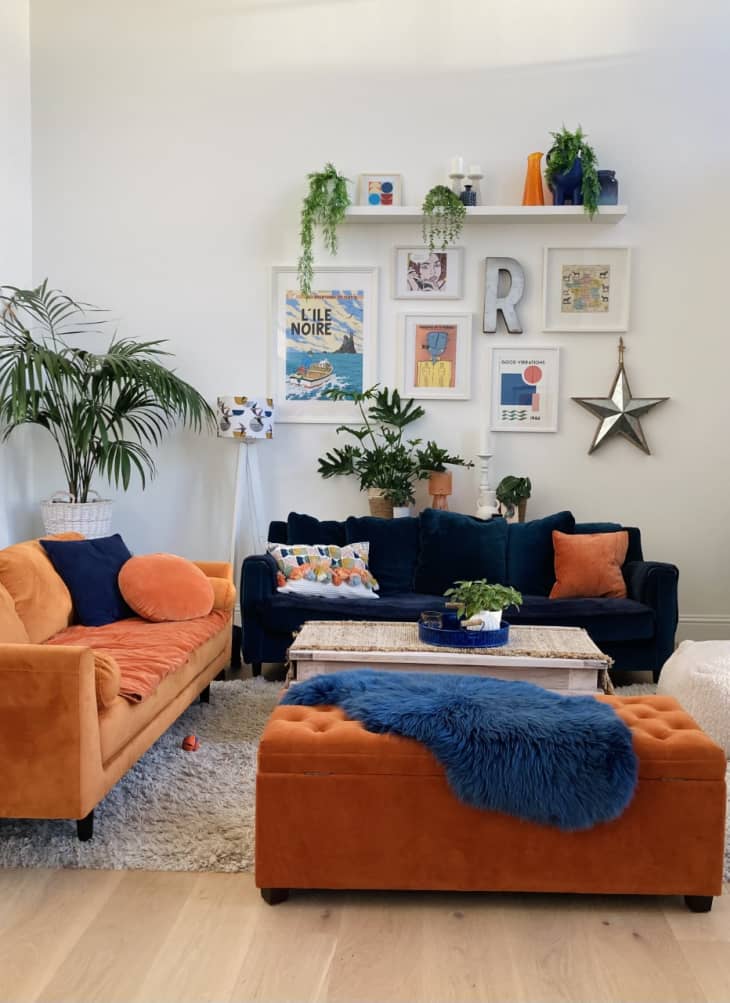 Living room with blue and orange sofas and an orange ottoman and a wall with lots of art work and plants.