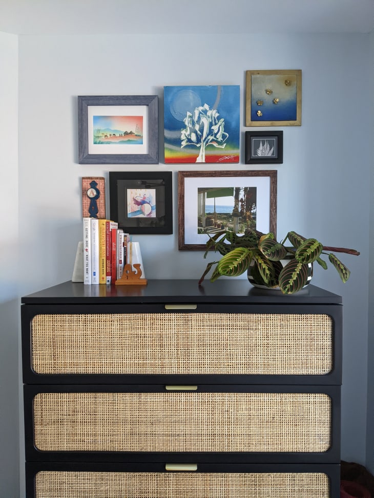 Black caned dresser with plants and books on top. Lots of framed artwork on the walls.