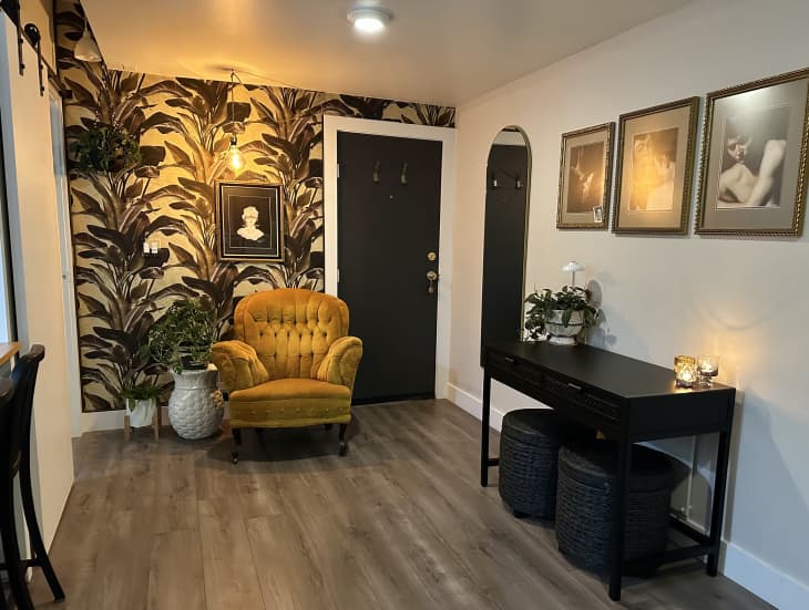 Apartment with lots of neutral, natural hues, Italian art, natural and black accents, plant=patterned wallpaper. This is a seating  area with a gold plush chair, black accent table with 2 black ottomans