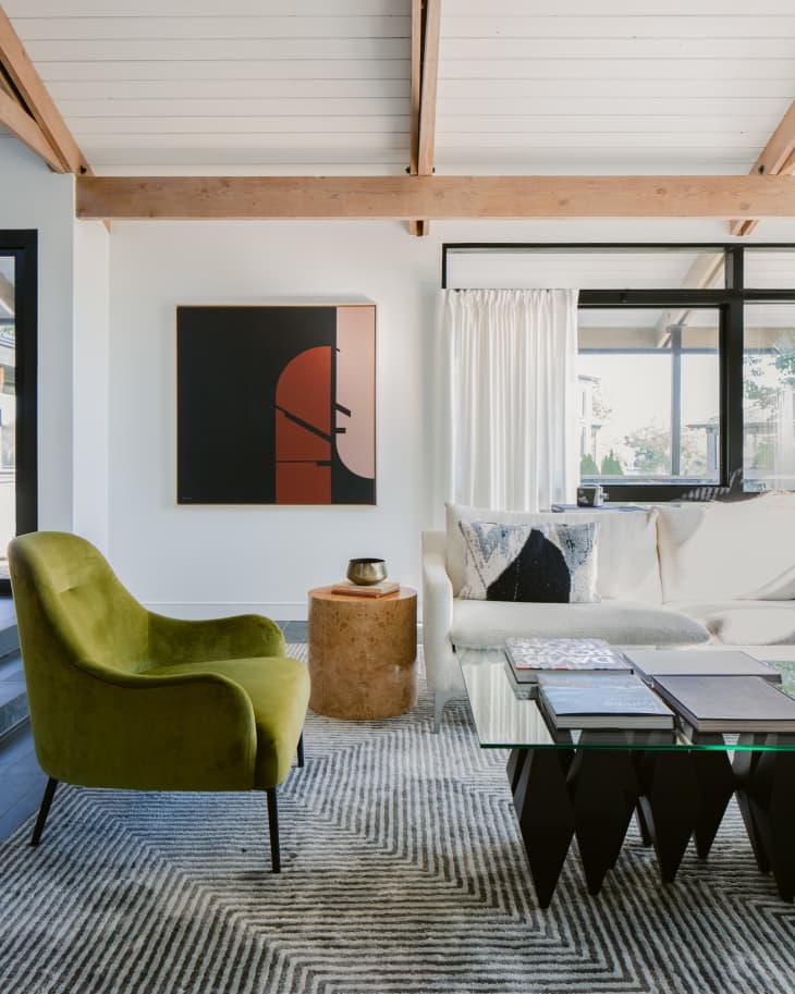 Contemporary living room with large white couch, green side chairs, glass and black coffee table, geometric black and white rug, and modern abstract wall painting, in a room with white ceilings, natural wood rafters, and large windows overlooking a lake.