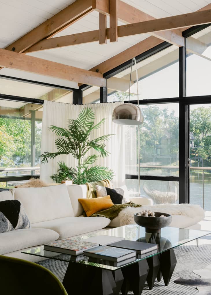 Contemporary living room corner with large white couch, glass and black coffee table, silver arched floor lamp, large palm plant, in a room with white ceilings, natural wood rafters, and large windows overlooking a lake.