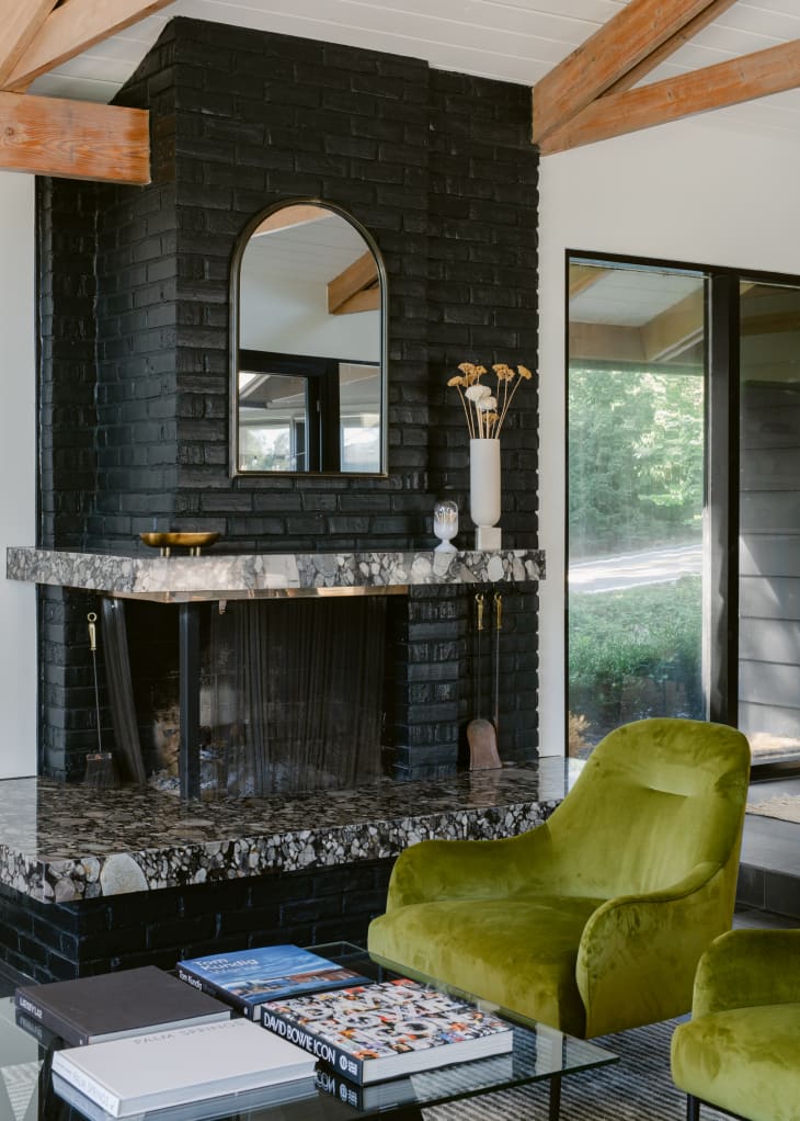 Pair of green velvet armchairs in front of black mid-century modern fireplace with black painted bricks, black and white marble fireplace surround and mantel, and arched mirror hanging on the brick.