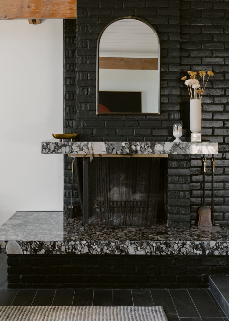 All black mid-century modern fireplace with black painted bricks, black and white marble fireplace surround and mantel, and arched mirror hanging on the brick.