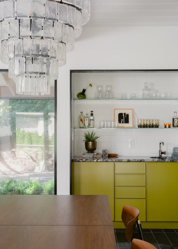 Mid-century modern dining room with glamorous crystal chandelier over a modern wood table, with a modern inset bar area with white tiles, glass shelves, and chartreuse-colored built-in cabinets.
