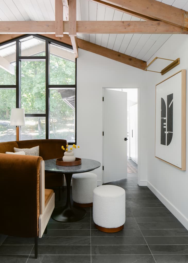 Kitchen breakfast nook with a round, rust-colored velvet dining banquette, round black table, two white leather poufs, and two tall wood lamps. Next to a large window with staggered black window trim, with black wood floors, white ceiling, and natural wood rafters.