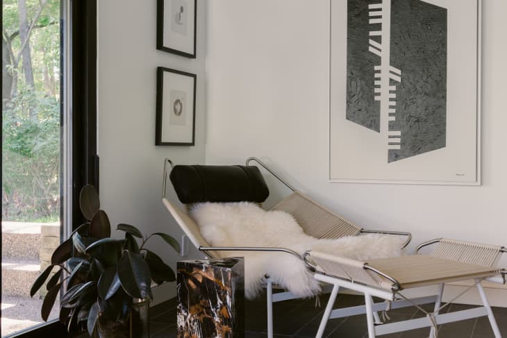 Corner of a room with modern fiber and metal armchair and ottoman, with a marble triangle side table, rubber leaf plant, and a black and white painting hanging on the wall created by artist John Neary.