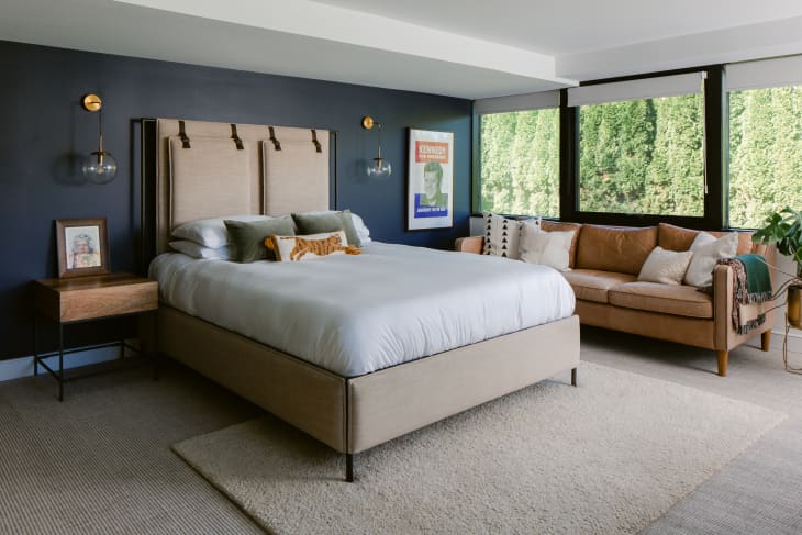 Modern bedroom with large windows, brown leather sofa, upholstered bedframe, and dark blue accent wall behind the bed.