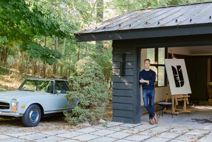 Artist John Neary standing in the corner of a MCM garage with the door open, a work-in-progress painting an easel inside, and a vintage light-blue car parked beside the garage