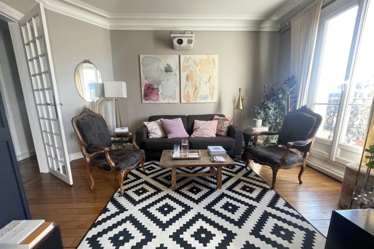 living room with black sofa, 2 black louis chairs, rustic wood coffee table, geometric pattern black and white rug, large windows with tall white curtains, muted watercolor artwork over sofa