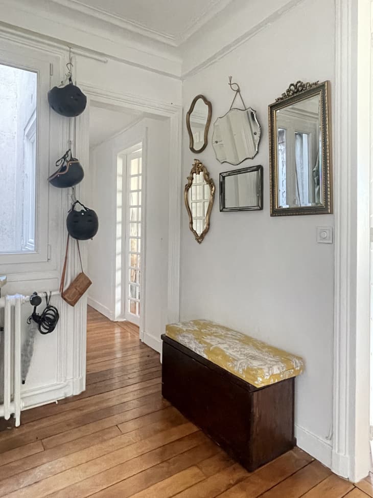 Paned window door open to hallway. White walls, wood floors, multiple framed mirrors of different shapes on the wall, pole with purses and bags hanging off. Bench underneath mirrors