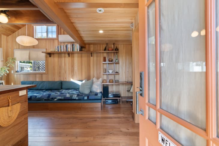 Exterior door opened to living space of beach cottage with long blue quilted sofa, Noguchi pendant lamp and open shelving displaying decorative objects.