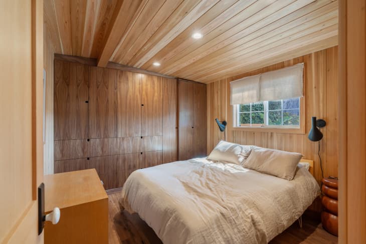 Bedroom with wood paneled walls and storage behind wood panels. Bed made with neutral colored bed linens.