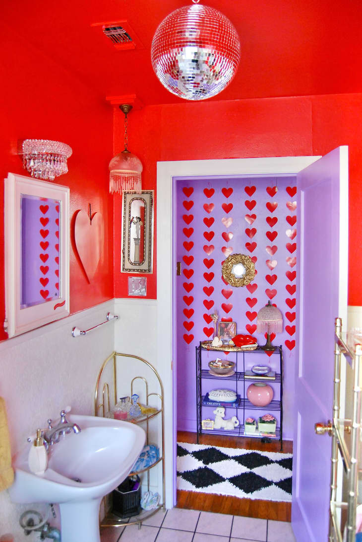 Bathroom with top half of wall painted bright red, the bottom half white. Framed art, crystals on light fixtures, delicate metal shelves. Lavender door and view into lavender hallway with red heart wall, black and white rug