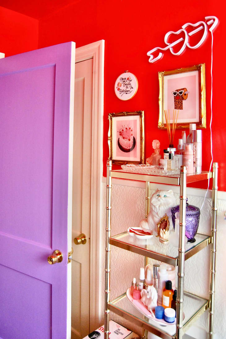Bathroom with top half of wall painted bright red, the bottom half white. Framed art, crystals on light fixtures, delicate metal shelves. Lavender door Bathroom with top half of wall painted bright red, the bottom half white. Framed art, crystals on light fixtures, delicate metal shelves. Detail of shelves, art, mirror, neon hearts on wall. Lavender door