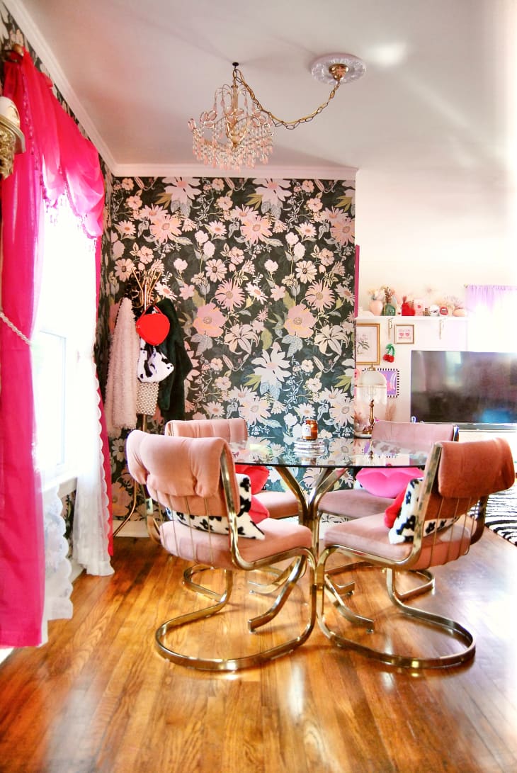 dining room with some pink walls, large window with bright pink/red curtains, one floral wallpaper wall, chandelier, pink velvet dining chairs with black and white cushions, tall gold deco bar cart