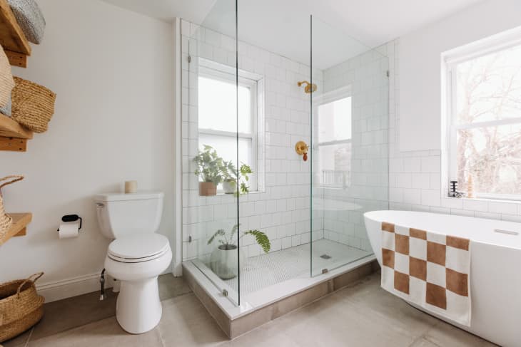 Philadelphia residence with white walls, lots of wood details: bathroom with oval freestanding tub, brown and white checkered bath mat, glass shower with white tiles, rose gold hardware, plants. Window over tub and in shower
