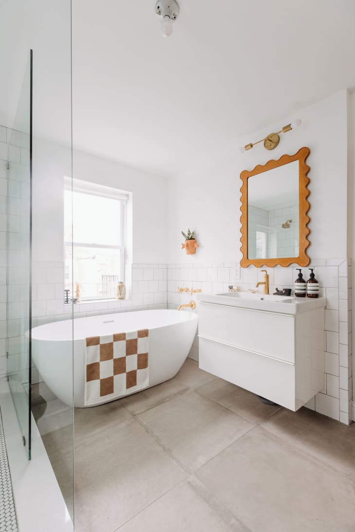 Philadelphia residence with white walls, lots of wood details: bathroom with oval freestanding tub, brown and white checkered bath mat, glass shower with white tiles, rose gold hardware, plants. Window over tub, white sink and cabinet with wavy frame mirror