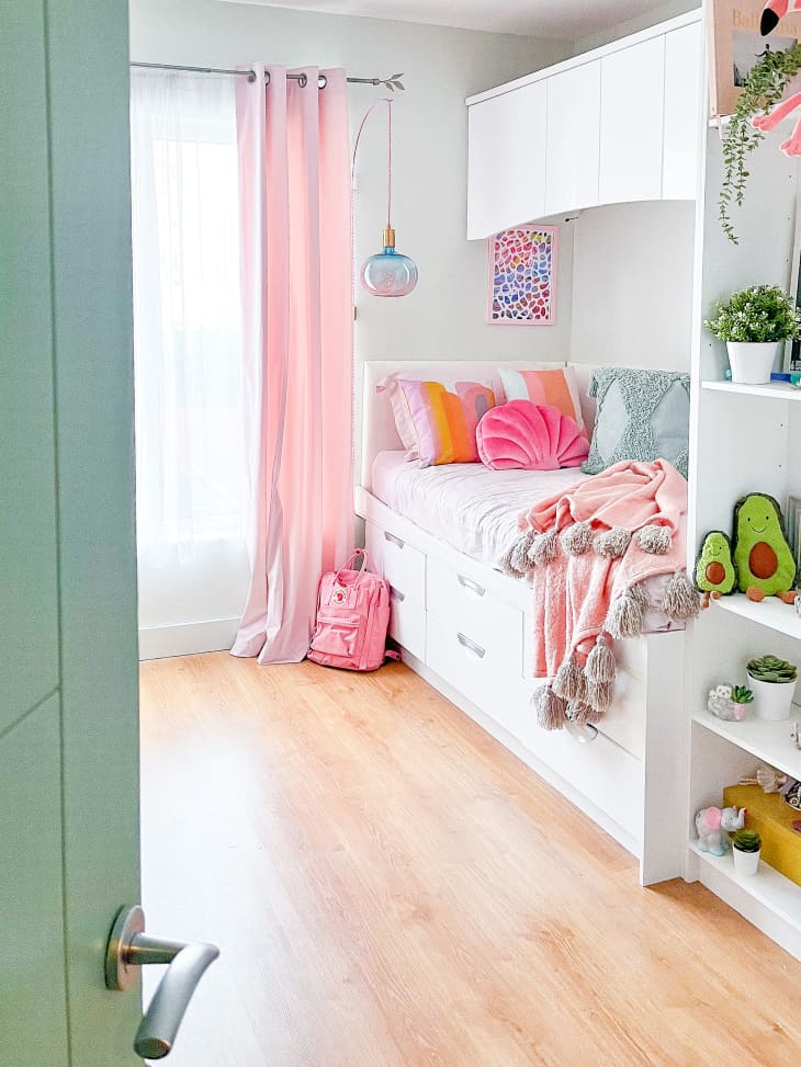 Kids bedroom with white bed with drawers, pink and orange throw pillows, pink throw blanket, light wood floor, white floor-to-ceiling shelving, pink curtains