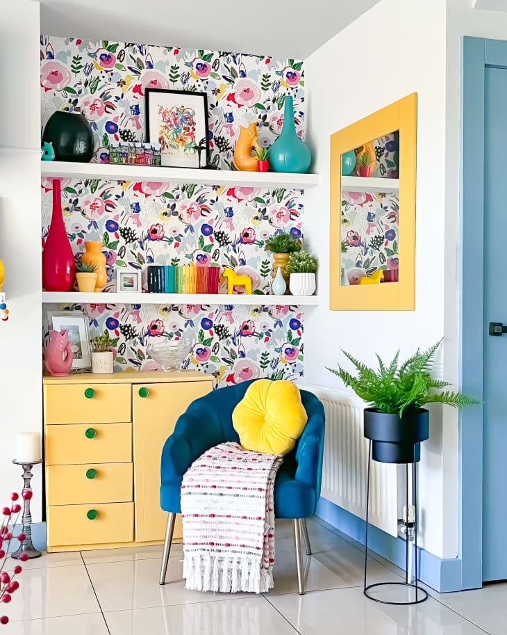 One side of living room with botanical wallpaper on one wall. White shelves with art objects, blue velvet accent chair with yellow pillow, plants around