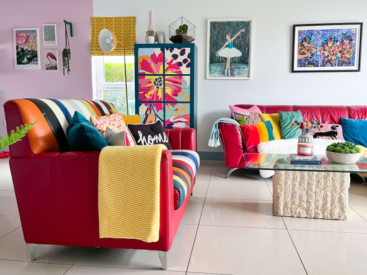 living room with red sofa and loveseat, colorful throw pillows, painted blue cabinet with flowers, glass coffee table with white base, colorful accents and art