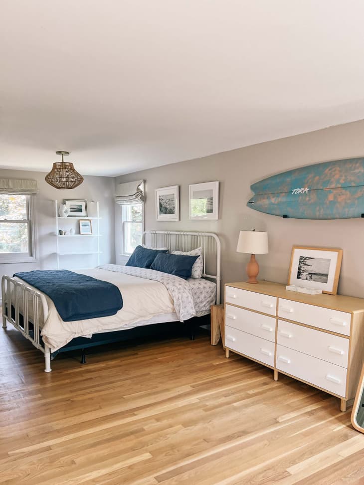Surfboard mounted above white dresser in newly renovated bedroom with white wrought iron bed frame and woven light fixture.