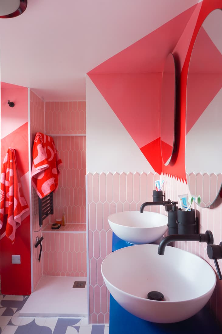 Ensuite bathroom after renovation. Walls have geometric designs painted in red, pink and white. There are blue sink cabinets, two white basin sinks with black hardware. Each sink has an oval mirror above. There is a walk in shower with a bench, all done in pink long hexagonal tiles with white grout. There are red and pink graphic designed colored towels