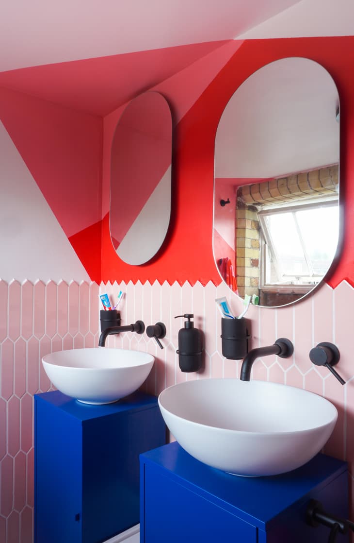Ensuite bathroom after renovation. Walls have geometric designs painted in red, pink and white. There are blue sink cabinets, two white basin sinks with black hardware. Each sink has an oval mirror above. Bottom half of walls done in pink long hexagonal tiles with white grout.