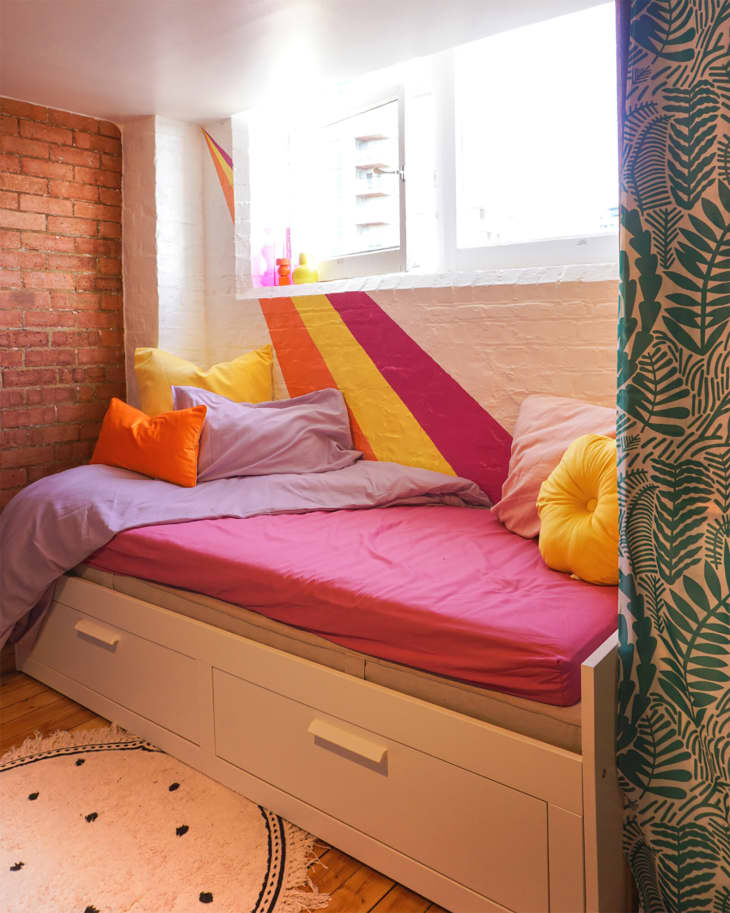 Bedroom after makeover/renovation. painted brick and exposed brick walls, daybed with storage and bright colored linens in shades of pink, orange, lilac, yellow. Hand painted wall stripes in pink, yellow, and orange.