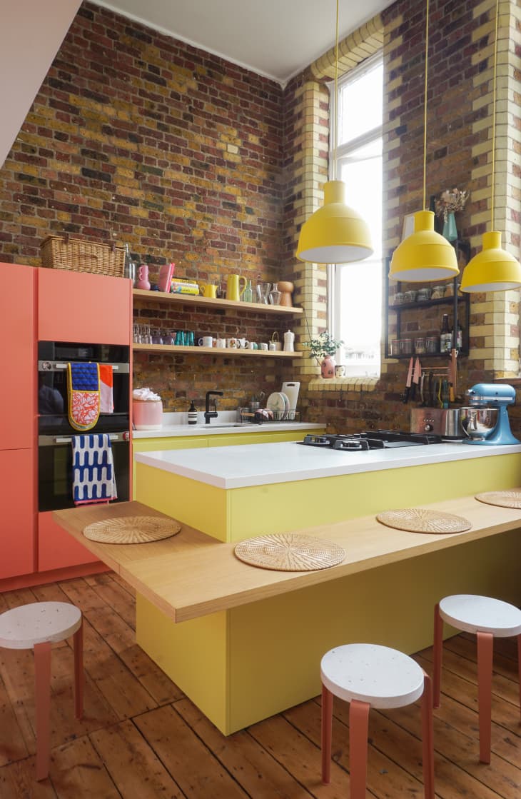 Kitchen with lots of exposed brick with different colored brick details around large windows, yellow cabinets, yellow pendant lights, coral pantry, wood bar projecting off island with coral and white barstools