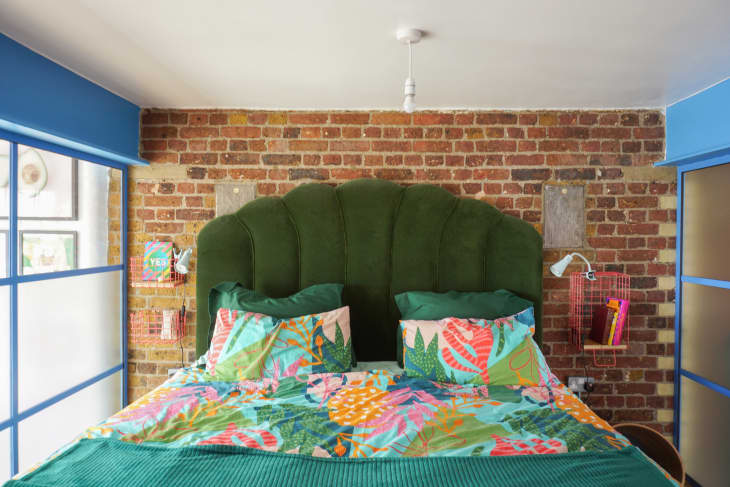 Main bedroom after being renovated. Exposed brick wall behind bed. Bed has olive green quilted velvet headboard, bedding is bright teal green with a colorful botanical print. There are blue trim/paned windows on either side. Living room gallery wall can be seen through window on left