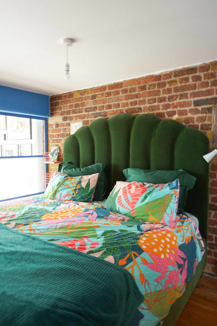 Main bedroom after being renovated. Exposed brick wall behind bed. Bed has olive green quilted velvet headboard, bedding is bright teal green with a colorful botanical print. There are blue trim/paned windows on either side. Living room gallery wall can be seen through window on left